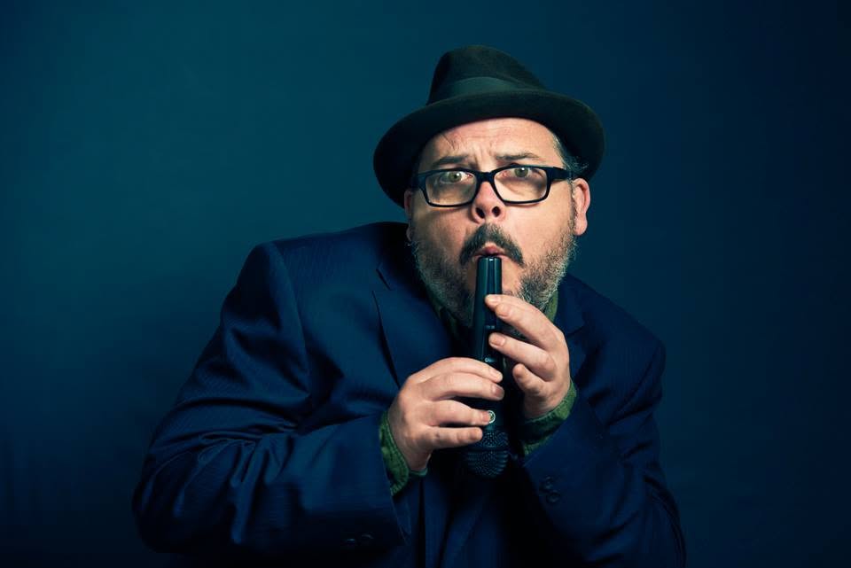 Tommy, in his black hat, leaning towards the microphone with a playful expression against a blue background.