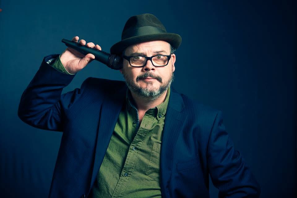 Tommy in his black hat and blue jacket, holding a microphone beside his head against a deep blue background.