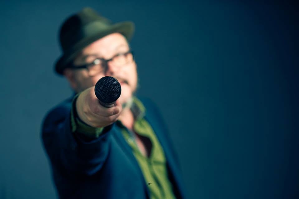 Tommy offers the microphone towards you, with his hat and blue backdrop behind it.